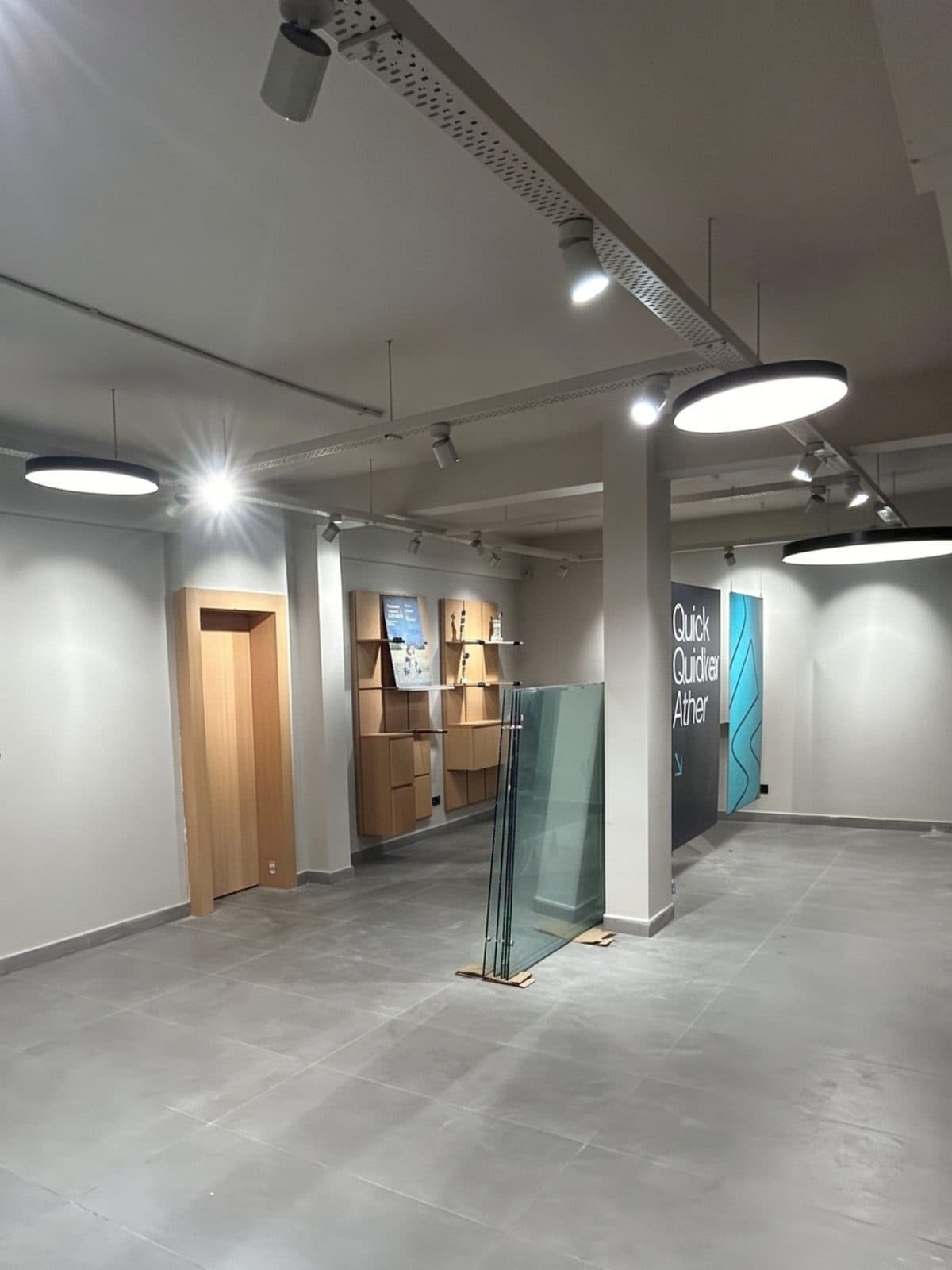 An experience centre part-way through fit-out: track lighting and circular pendants installed, oak display joinery fixed to the wall, and glass panels still standing against a column waiting to be set.