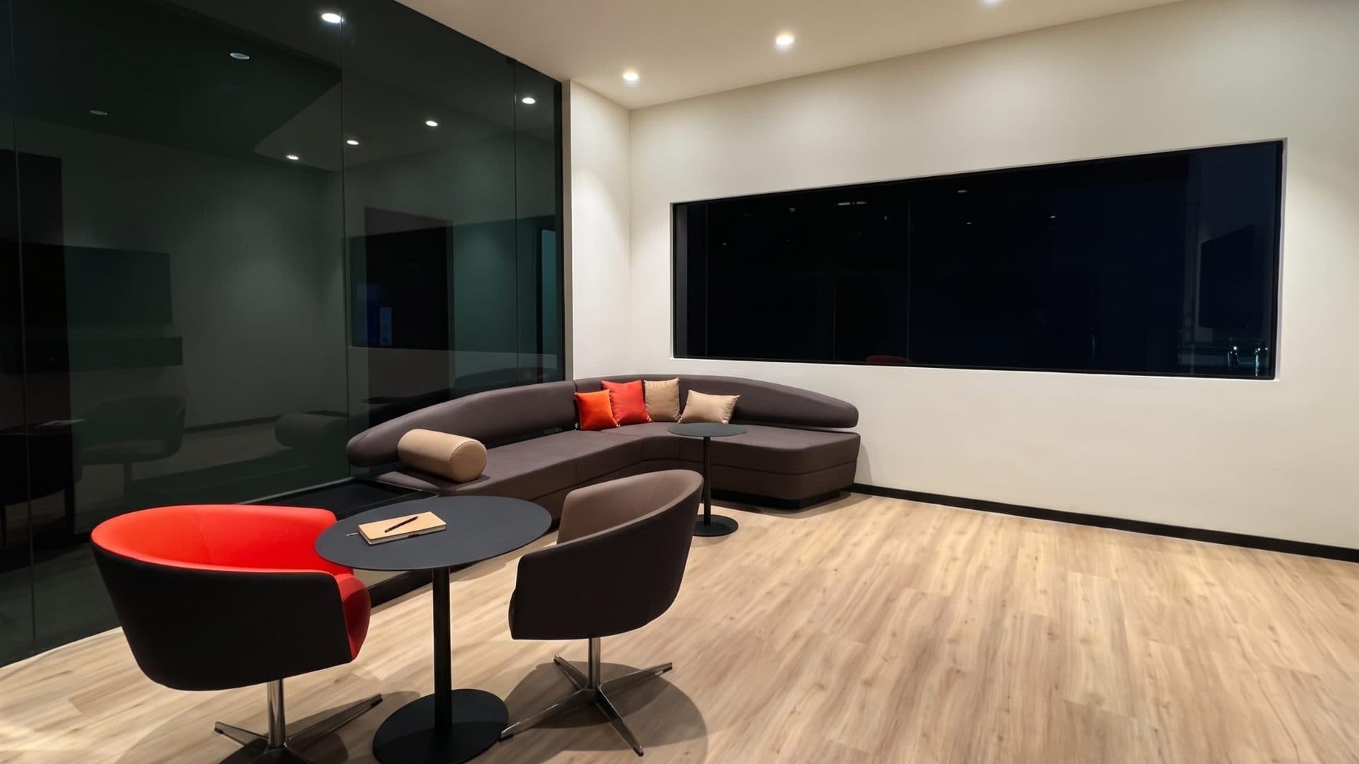 A customer lounge with a curved sofa, low tables and tub chairs in red and brown, on a pale oak floor beside a glazed partition.