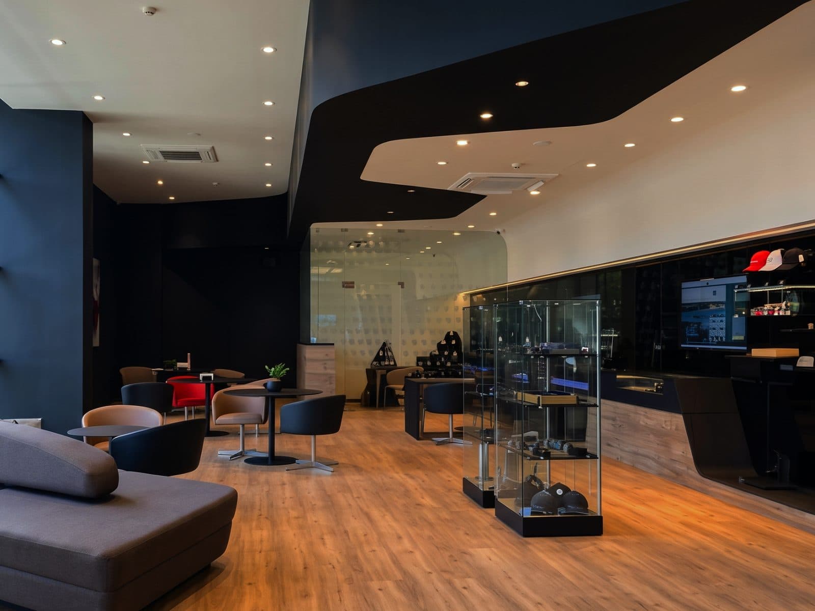 A showroom lounge under a sculpted white and black ceiling, with a glass merchandise cabinet and low seating on an oak floor.